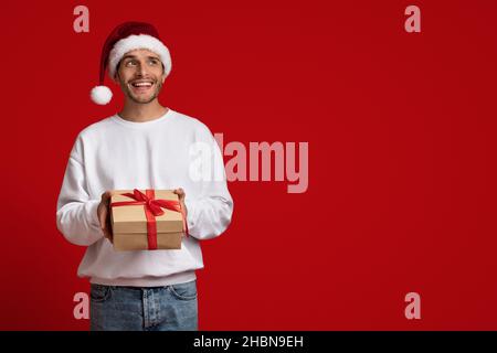 Dreamy Young Man In Santa Hat Holding Gift Box And Looking Away Stock Photo