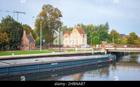 Freight ship sails through a canal near Dampoort gate in Bruges, Belgium. Picturesque lock keeper's cottages line the waterfront. Stock Photo