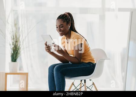Pretty black lady watching movie, using wireless earphones and tablet pc while sitting on chait at home Stock Photo