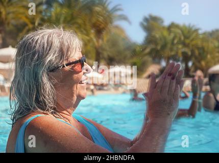 Elderly senior woman with grey hair, wearing blue swimsuit doing water aerobics in hotel pool, clapping her hands Stock Photo
