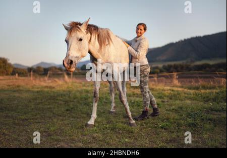 White Arabian horse, autumn afternoon, detail on head, blurred smiling young woman in warm jacket holding its back Stock Photo