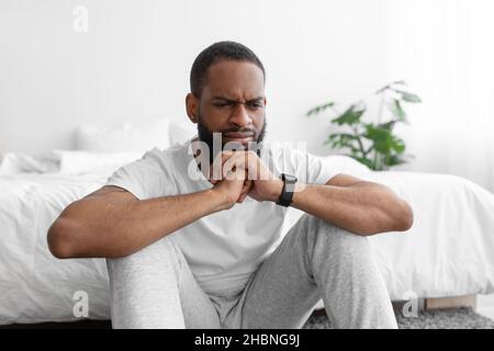 Pensive upset young black bearded man in white clothes sits on floor, near bed, ponders Stock Photo
