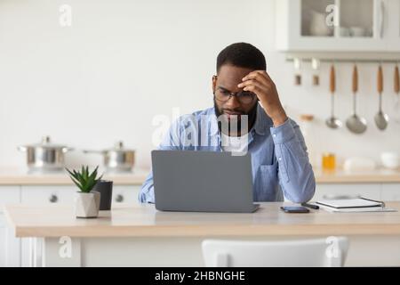 Sad tired pensive young black bearded guy in glasses works at laptop, ponders problem solution in scandinavian kitchen Stock Photo