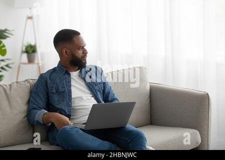 Serious concentrated young black bearded guy work on laptop on sofa in room interior, look at window Stock Photo