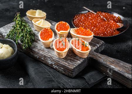 Tartlets with Red caviar, butter and thyme on wooden board. Black background. Top view Stock Photo