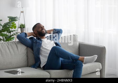 Happy satisfied relaxed young black bearded male resting in living room interior with tablet on sofa Stock Photo