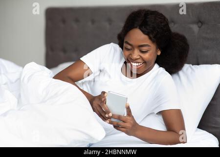 Smiling black lady using cellphone while laying in bed Stock Photo
