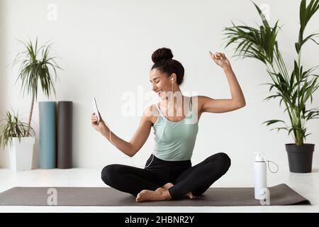 Wellbeing Concept. Cheerful Young Woman Sitting On Yoga Mat And Listening Music Stock Photo