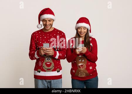Online Communication. Couple In Santa Hats And Christmas Sweaters Messaging On Smartphones Stock Photo