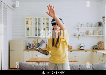 Positive young woman in casual clothes with modern bionic arm and virtual reality headset dances near sofa in stylish kitchen Stock Photo