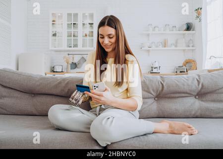 Pretty happy woman with prosthesis arm pays for online order with credit card via mobile phone on sofa in living room closeup Stock Photo