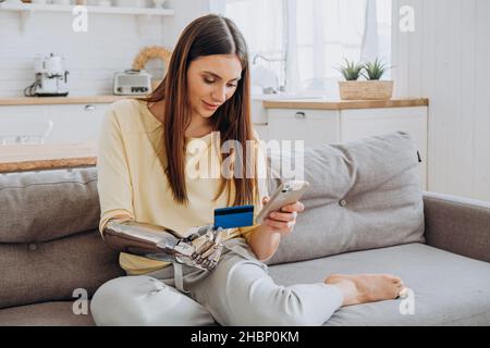 Pretty happy woman with prosthesis arm pays for online order with credit card via mobile phone on sofa in living room closeup Stock Photo