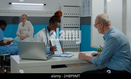 Senior patient looking at tablet with chiropractic injury while young doctor holding device and explaining osteopathy. Medic and old man at desk analyzing bone examination results Stock Photo
