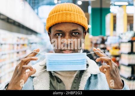 Young African-American man in stylish knitted hat puts on blue disposable mask standing in supermarket department close view Stock Photo