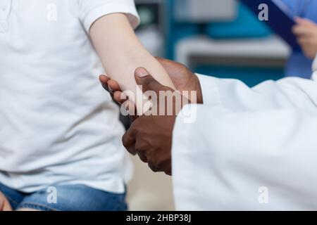 Closeup of african american pediatrician doctor rubbing injured arm of little patient during medical physiotherapy in hospital office. Young kid having broken bone after accident. Health care service Stock Photo