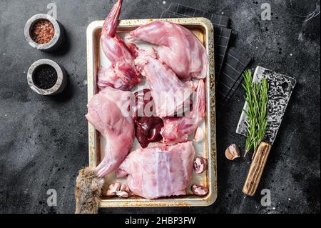 Raw wild hare slices legs, fresh game meat in steel tray with herb. Black background. Top view Stock Photo