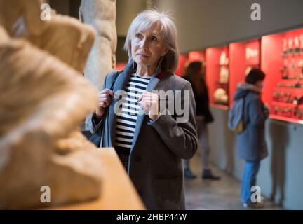 Interested elderly woman examining ancient sculpture in museum Stock Photo