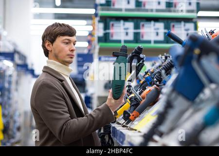 A young man carefully selects a perforator in a construction supermarket for repairs in an apartment, holds two electric perforators in his hands. Stock Photo