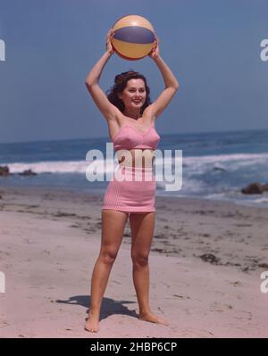 Young, dark haired woman in pink 2-piece bathing suit standing on the beach holding a beach ball above her head Stock Photo