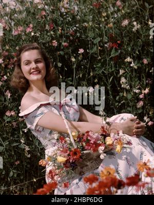 Young woman in white print Summer dress sitting holding a basket of flowers against a backdrop of a field of flowers Stock Photo