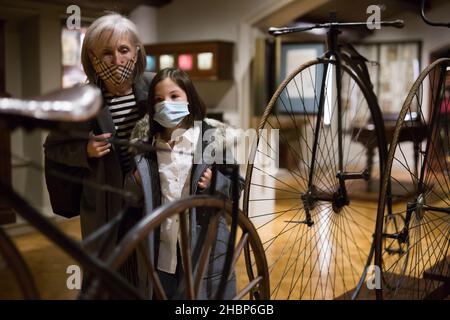Elderly woman with preteen granddaughter in protective masks examining retro bicycles in museum Stock Photo