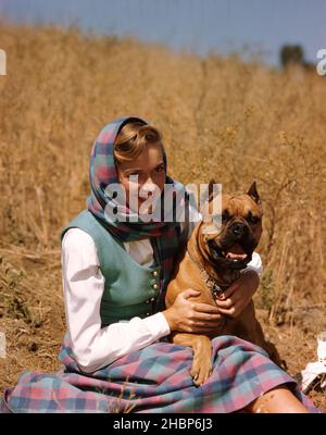 Young teen woman wearing a checkered skirt and matching head wrap sitting in a field with her Boxer dog Stock Photo