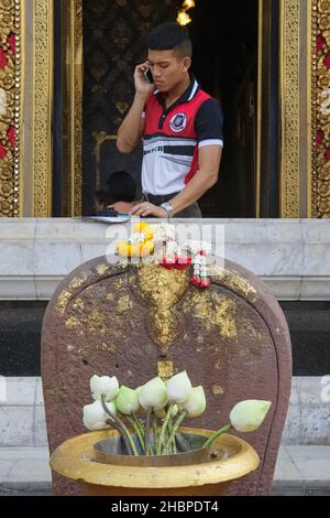 A man standing by a window at (temple) Wat Boworniwet in Bangkok, Thailand, is speaking on his mobile phone, ornamental lotus flowers in the f/g Stock Photo