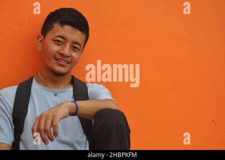 A happy Asian guy looking at camera, sitting against orange wall background with copy space Stock Photo