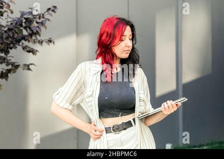 Portrait of a female student or businesswoman with creative red and black hair in smart casual clothing using digital tablet outdoors near the office Stock Photo