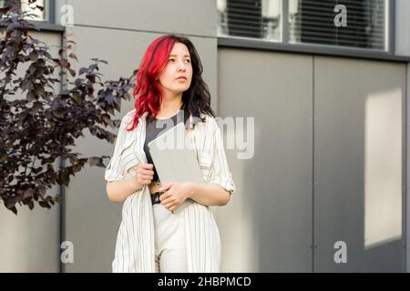 Portrait of a female student or businesswoman with creative red and black hair in smart casual clothing using digital tablet outdoors near the office Stock Photo