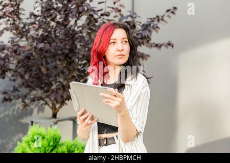 Portrait of a female student or businesswoman with creative red and black hair in smart casual clothing using digital tablet outdoors near the office Stock Photo