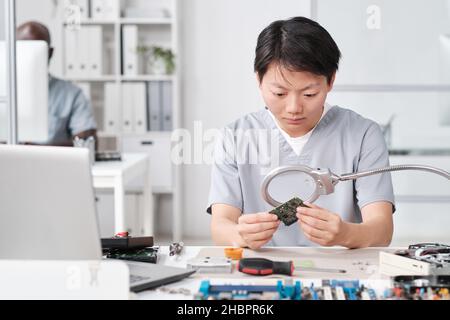 Young Chinese female in uniform looking at microchip in board through magnifying glass while sitting by desk Stock Photo