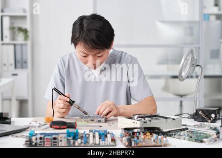 Young Chinese female technician in uniform repairing circuit board with microprocessor while sitting by workplace Stock Photo