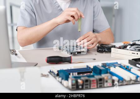 Hands of young repairperson in uniform fixing details of circuit board with screwdriver while sitting by desk in laboratory Stock Photo