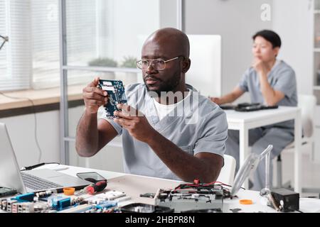 Young serious repairman looking at circuit board of electronic device in his hands while sitting by workplace against female co-worker Stock Photo