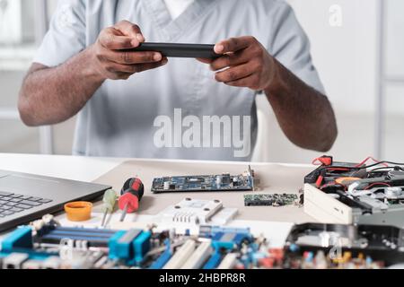 Hands of young African repairman photographing circuit board of electronic device while sitting by desk in workshop Stock Photo