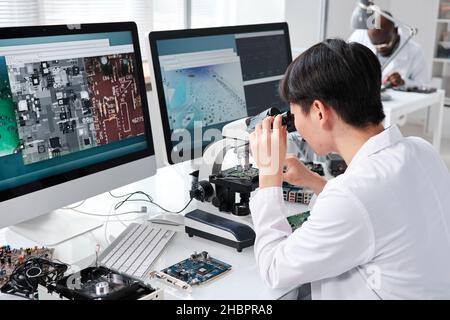 Young Asian female researcher in whitecoat looking in microscope while sitting by desk in front of computer monitors Stock Photo