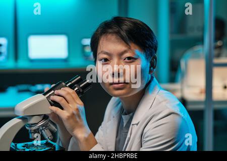 Young confident female scientist or technician with microscope looking at you while sitting by workplace in laboratory Stock Photo