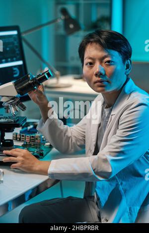 Young Chinese woman in whitecoat looking at camera while working with microscope in technical laboratory Stock Photo