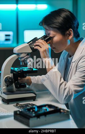 Young Chinese woman in whitecoat looking in microscope lens while studying microchip scheme in laboratory Stock Photo