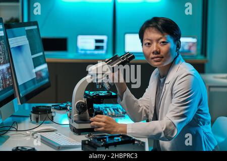 Young Asian researcher in whitecoat working with microscope and looking at you while sitting by workplace in laboratory Stock Photo