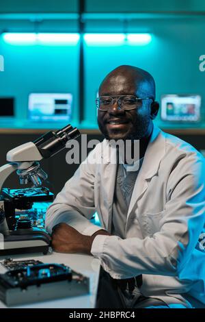 Happy young male laboratory worker in whitecoat and eyeglasses sitting by desk with microscope and looking at you with smile Stock Photo