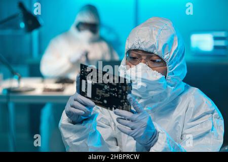 Young Asian female in protective workwear looking at circuit board in her gloved hands while working in laboratory Stock Photo