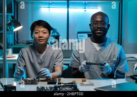 Two young successful technicians in gloves and uniform working by desk and looking at camera in laboratory or workshop Stock Photo