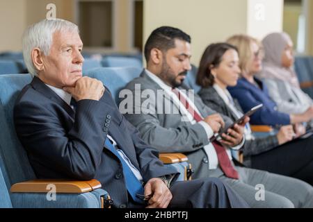 Senior businessman or politician in formalwear listening to speaker at conference while sitting in row with foreign colleagues Stock Photo