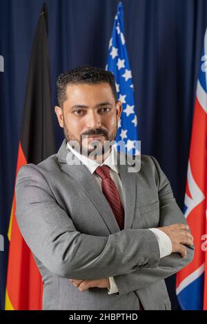 Serious mature businessman or politician crossing arms by chest while standing against row of flags in conference hall Stock Photo