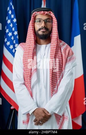 Young Arabian man in turban and national clothes keeping hands put together while standing in front of camera Stock Photo
