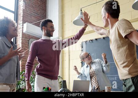 Two young successful businessmen giving each other high five among their happy intercultural female colleagues Stock Photo