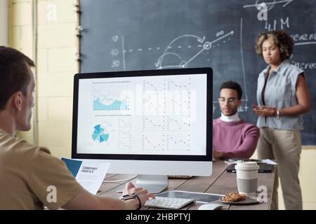 Young serious broker analyzing financial graphs on computer screen while sitting by table in front of two intercultural colleagues Stock Photo