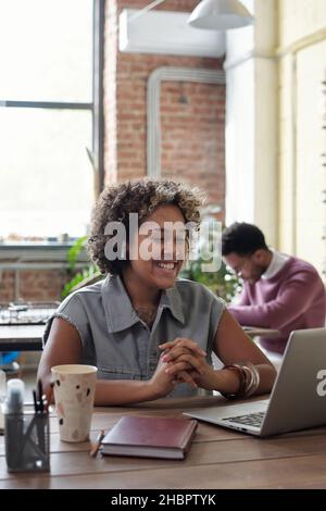 Happy young businesswoman with earphones watching online video while sitting by table in front of laptop against co-worker Stock Photo